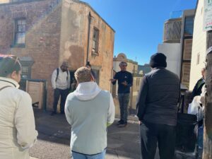tour guide with group who all have backs to the camera. Guide standing between buildings, including on his left an old brick building. 